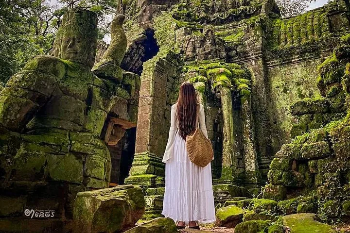 Mujer de pie frente a una antigua ruina de templo de piedra cubierta de musgo verde.